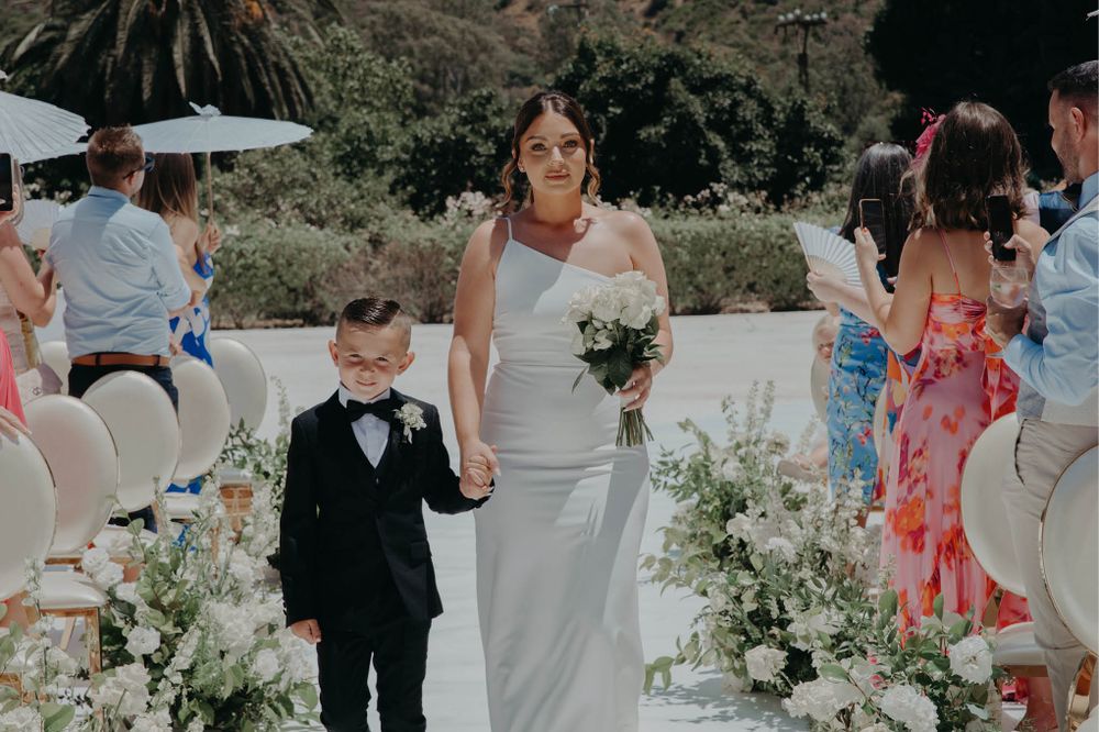 A young ring bearer in a tuxedo walks down the aisle with the bride during an outdoor wedding ceremony.