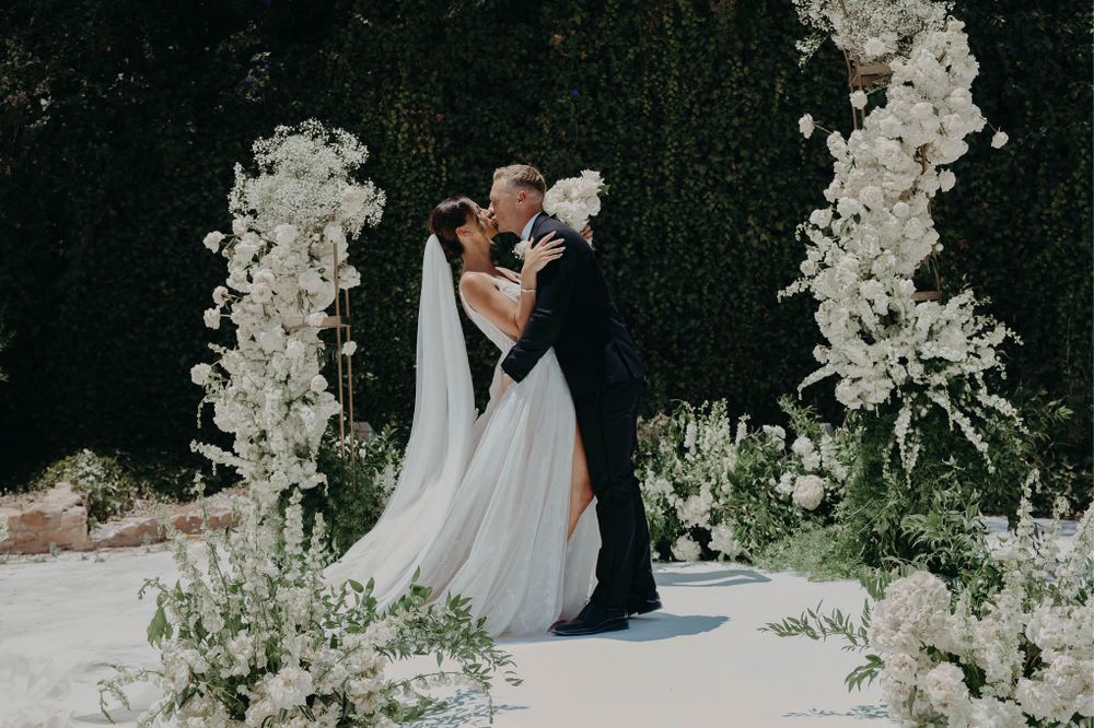 Newlyweds share a romantic moment surrounded by white floral arrangements against a dark hedge wall.
