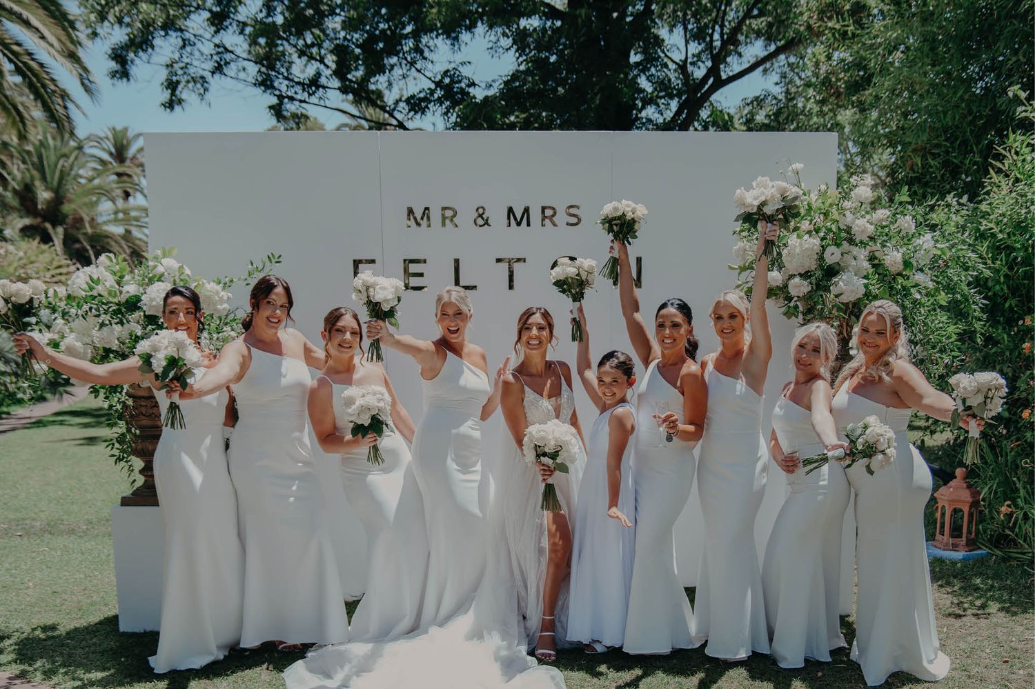 Bridesmaids in white dresses pose together in front of a Mr & Mrs sign at an outdoor wedding.