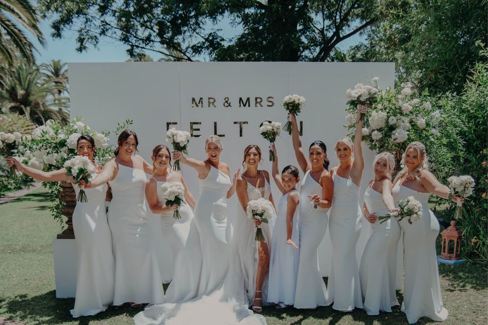 Bridesmaids in white dresses pose together in front of a Mr & Mrs sign at an outdoor wedding.