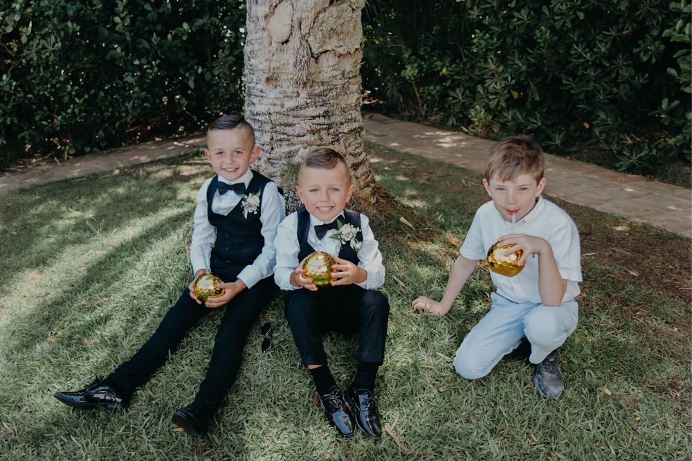 Three young boys in formal attire sit on grass enjoying snacks during a wedding reception.