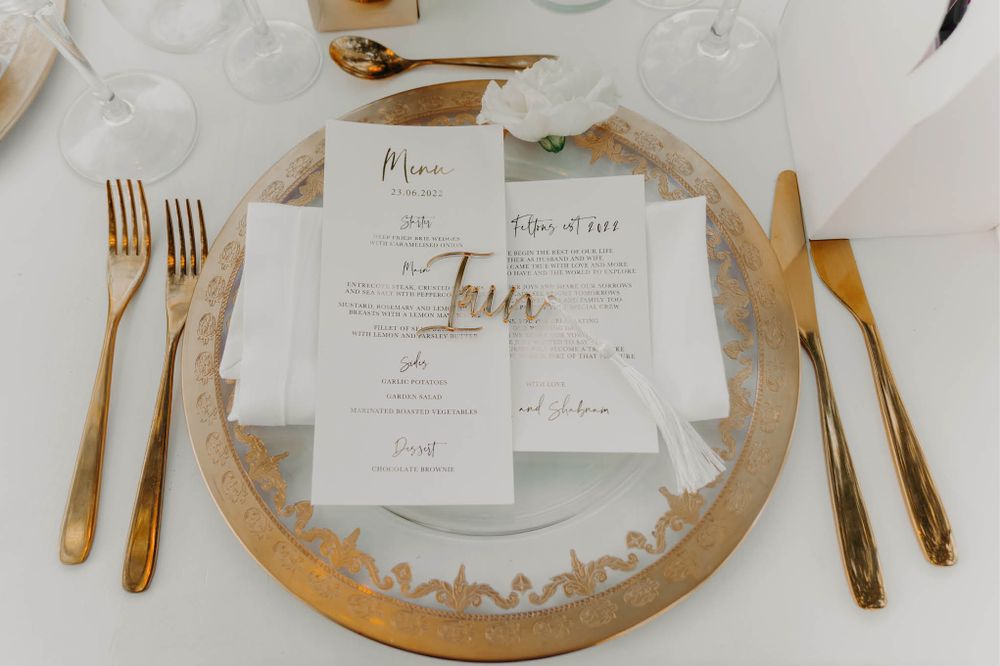 Wedding place setting with gold cutlery and menu card on decorative charger plate.