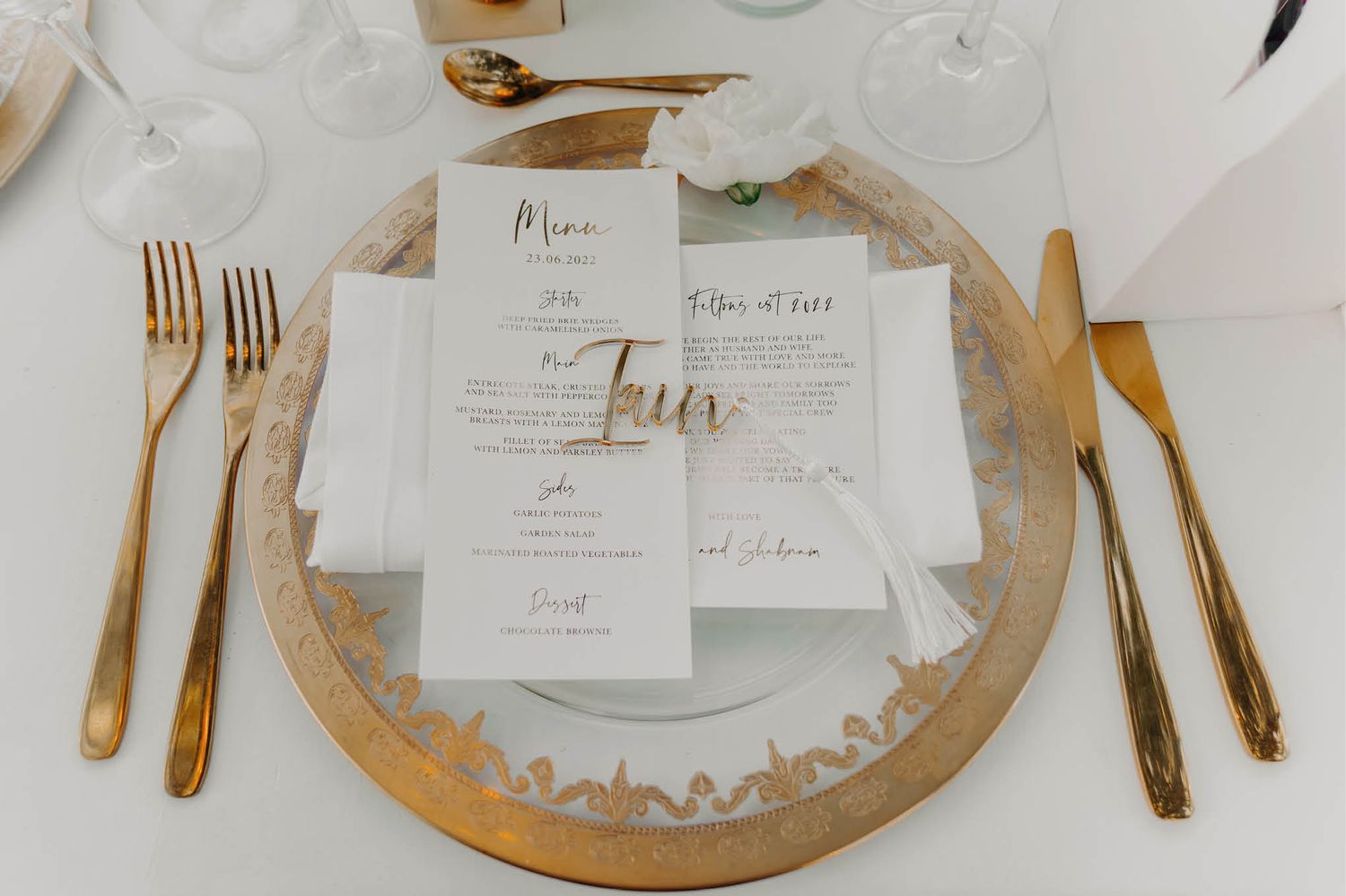 Wedding place setting with gold cutlery and menu card on decorative charger plate.