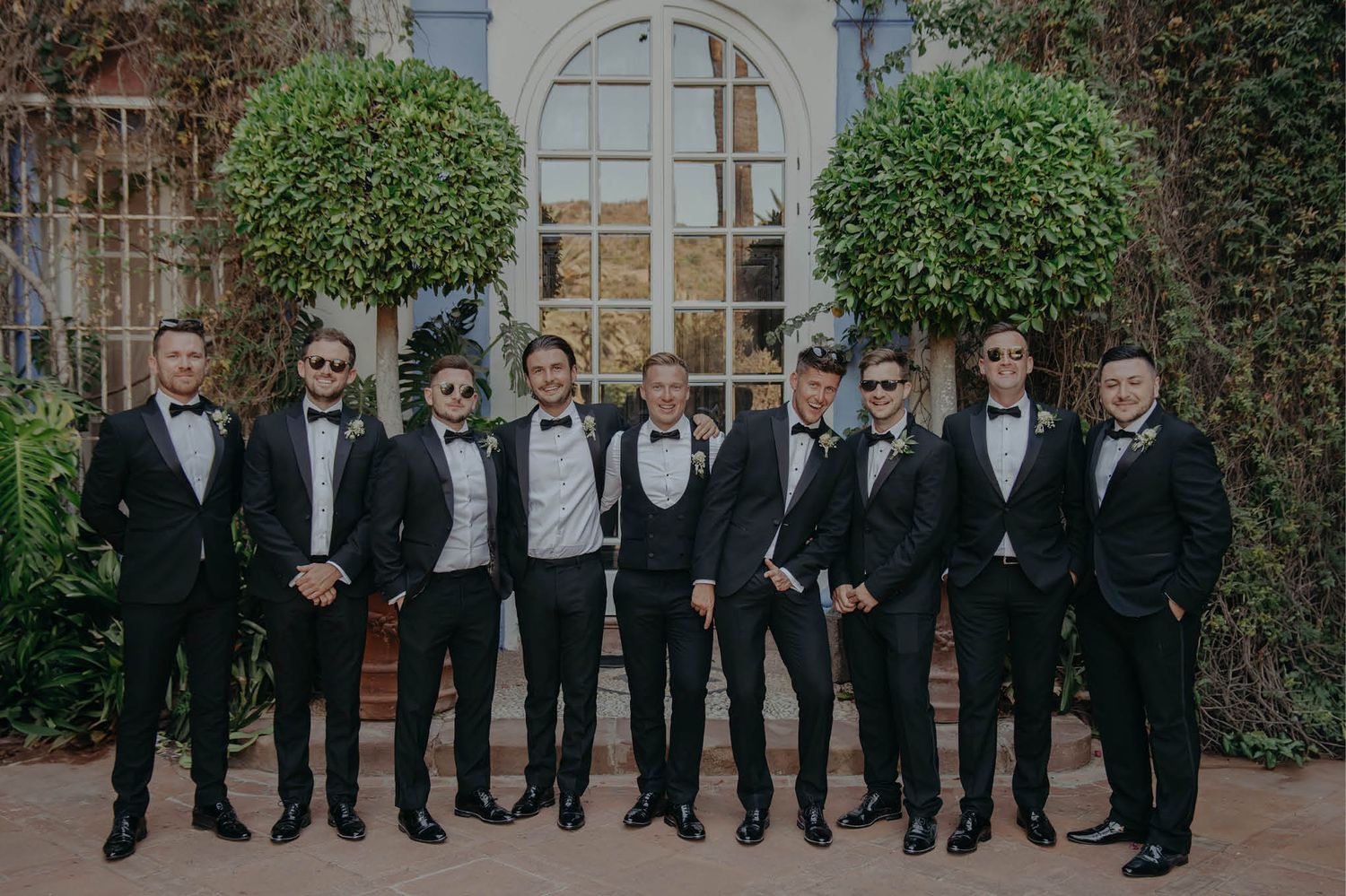 Wedding party members in formal black tuxedos pose in front of arched doorway.