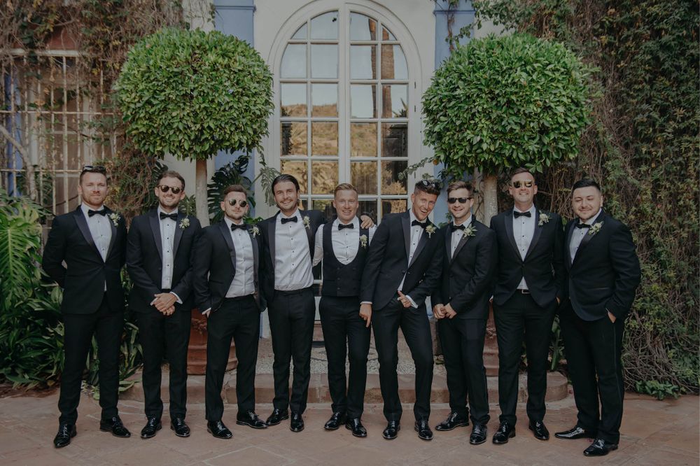 Wedding party members in formal black tuxedos pose in front of arched doorway.