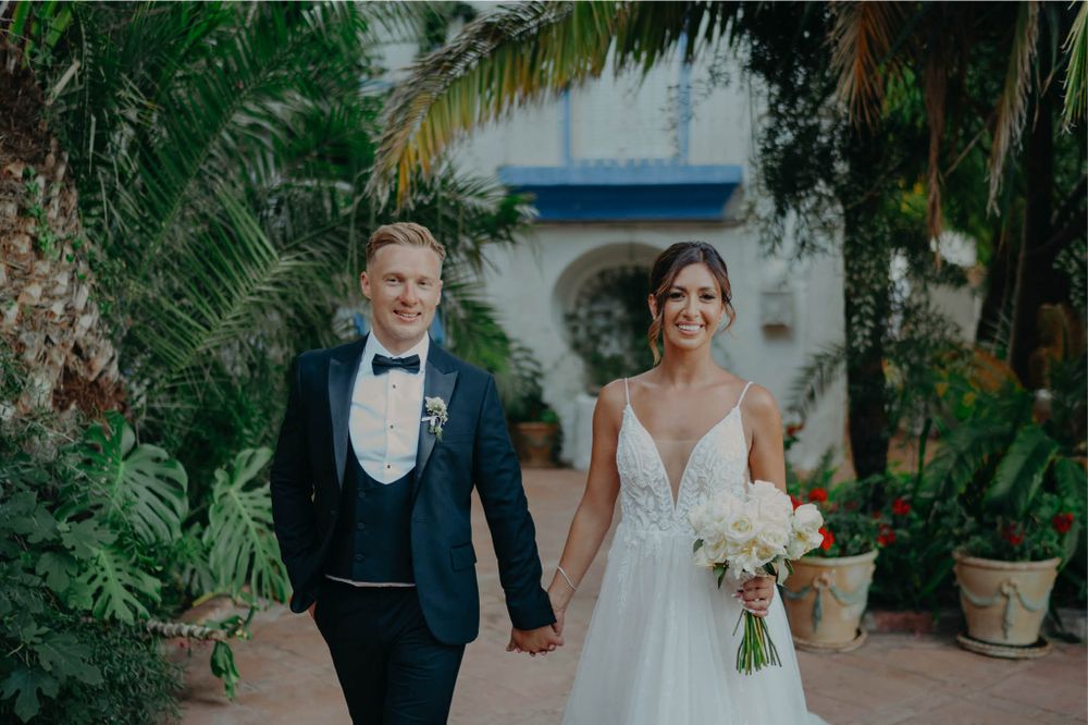 Wedding couple walks hand in hand through a lush garden path with palm trees and Mediterranean architecture.