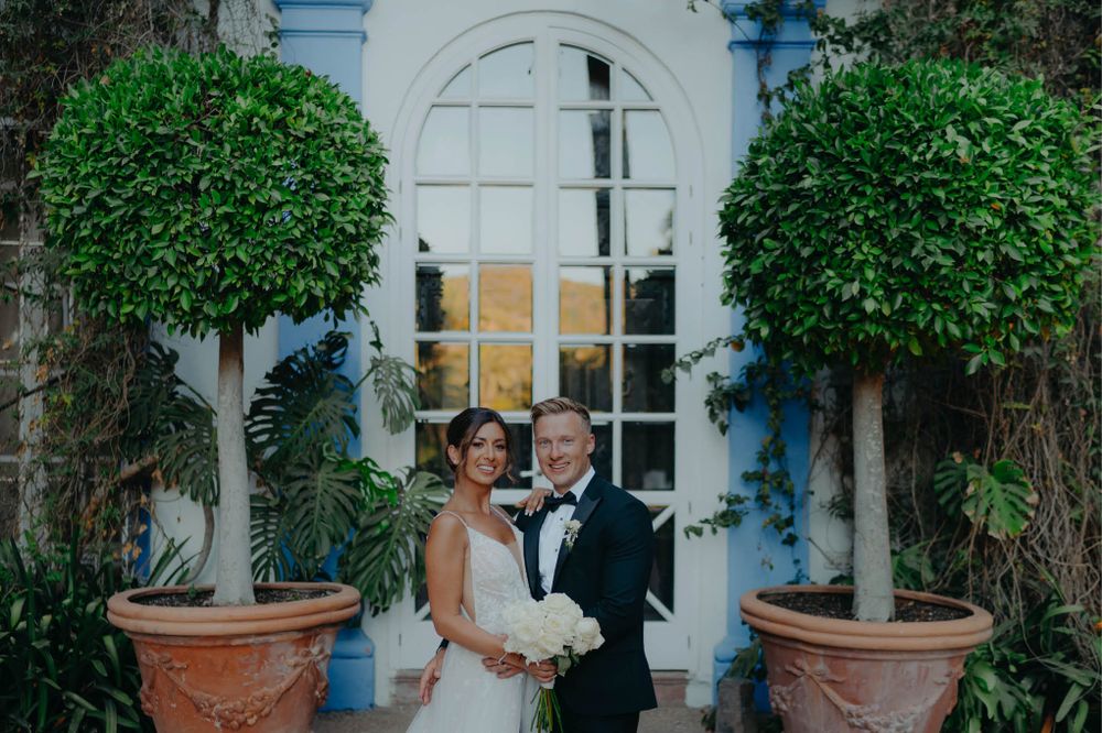 Wedding portrait in front of a beautiful white arched doorway flanked by manicured topiary trees.
