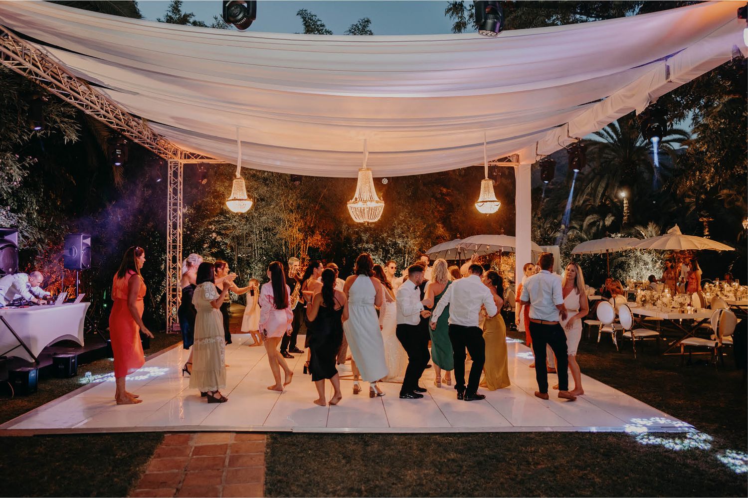 Wedding guests dancing under an elegant draped canopy with hanging lanterns at an outdoor evening reception.