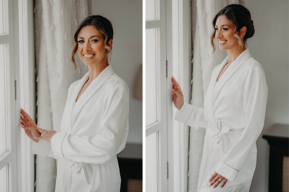 A woman in a white bathrobe poses elegantly by a window during wedding preparations.