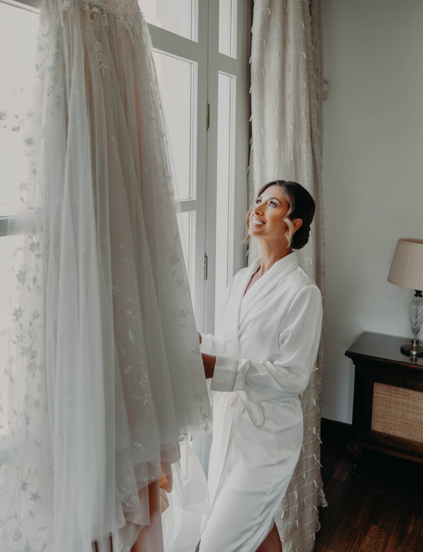 A white wedding dress hangs by a window as someone in a robe prepares for the special day.