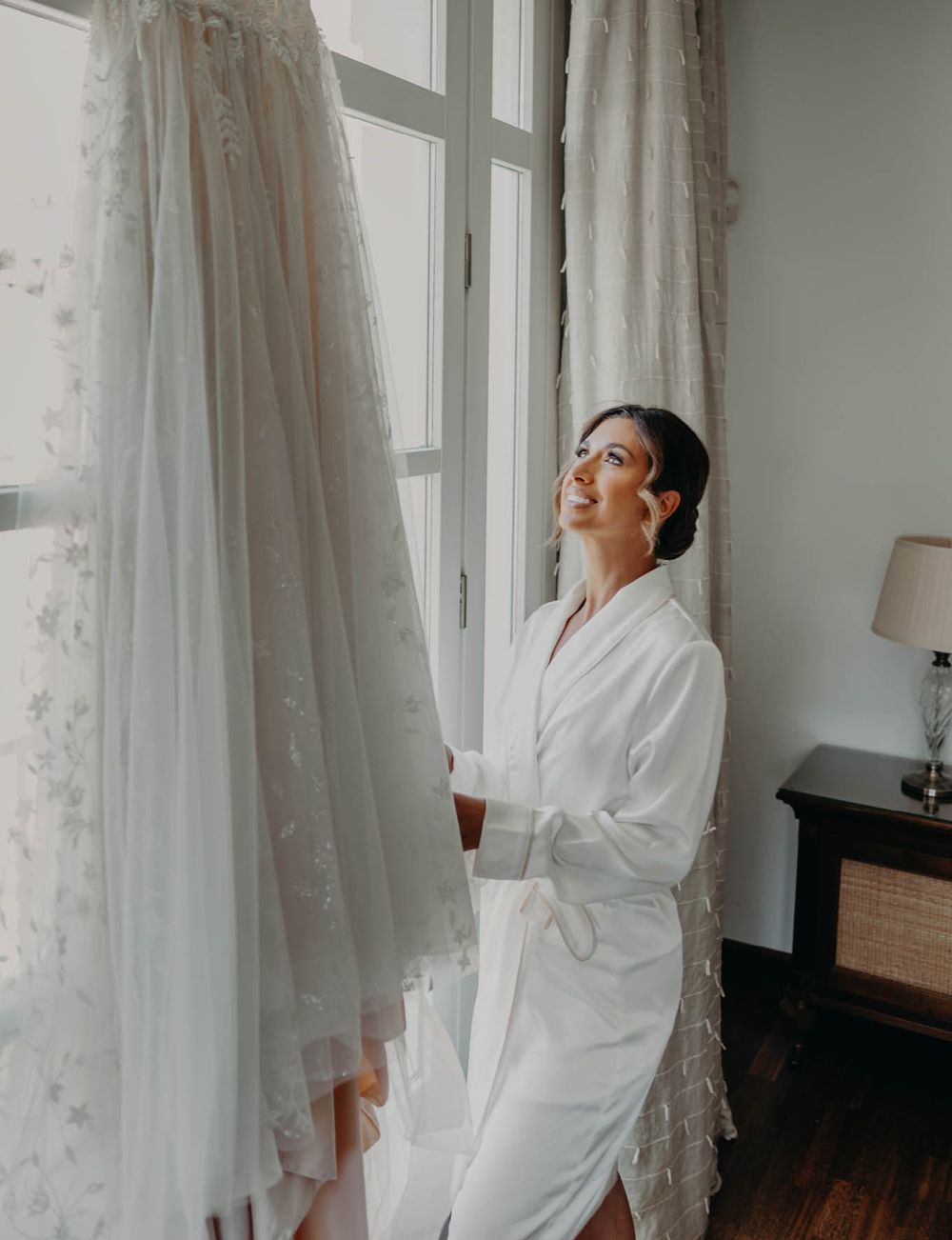A white wedding dress hangs by a window as someone in a robe prepares for the special day.