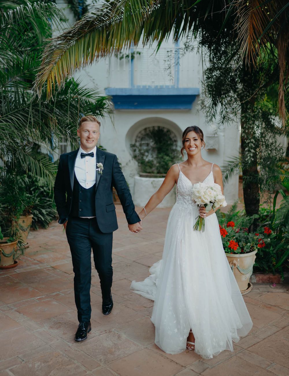 Wedding couple walking hand in hand along Mediterranean villa pathway with palm trees.