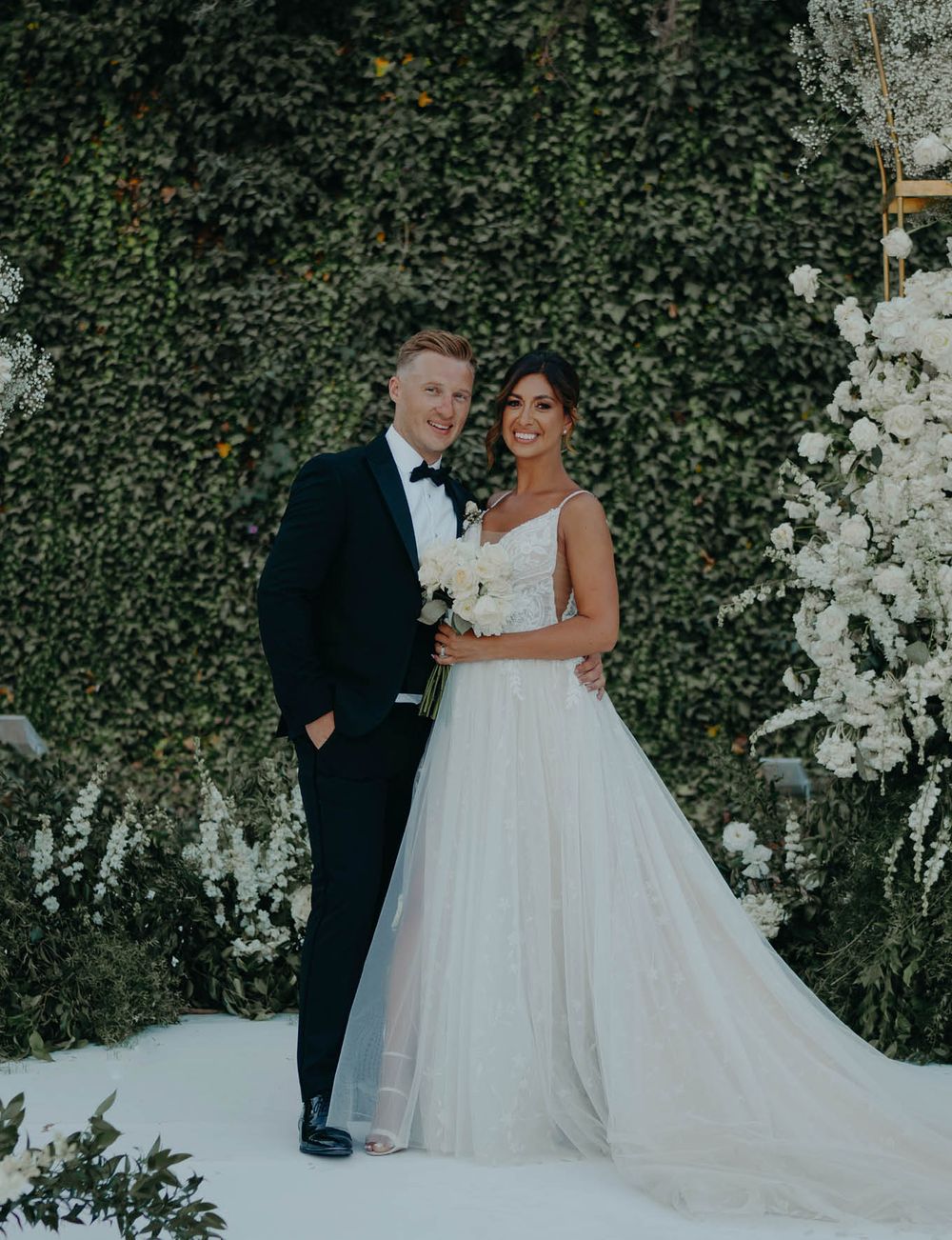 Elegant wedding portrait against white floral arch and ivy backdrop.