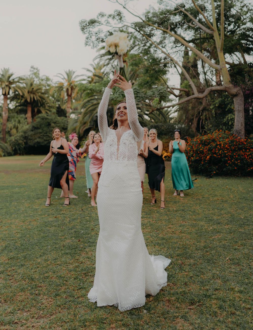 Bride in white lace gown tosses bouquet during outdoor garden ceremony.