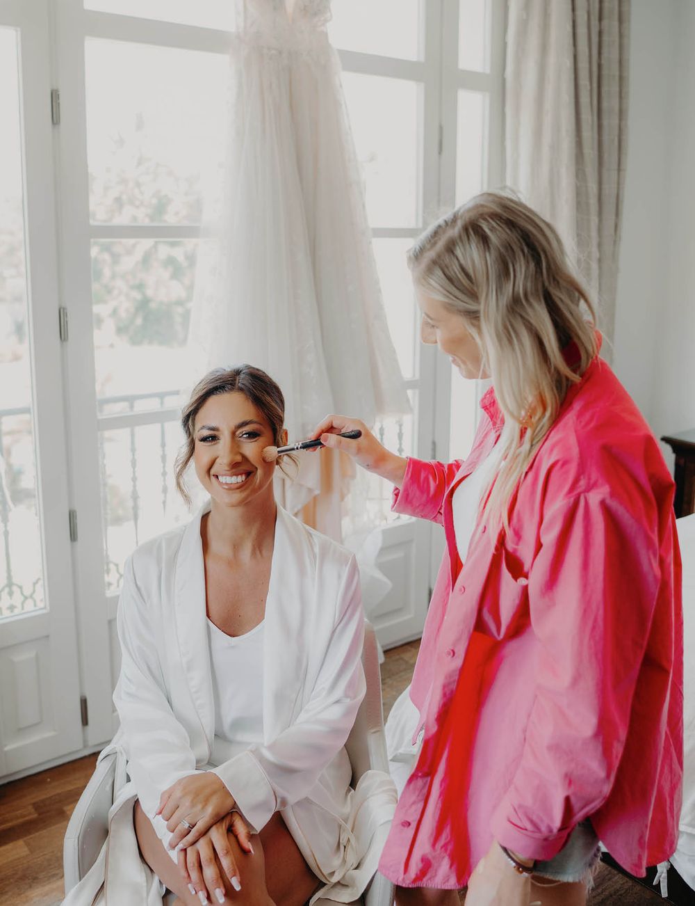 Getting ready scene showing makeup application in a bright room with a wedding dress hanging nearby.