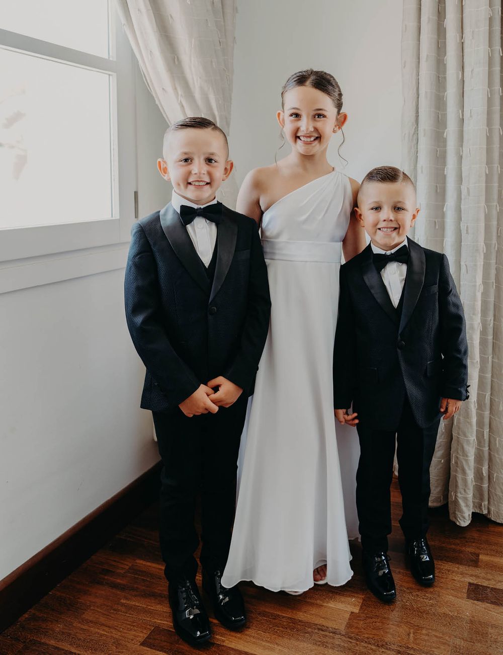 Three formal figures pose together by a window, creating a charming wedding day portrait.