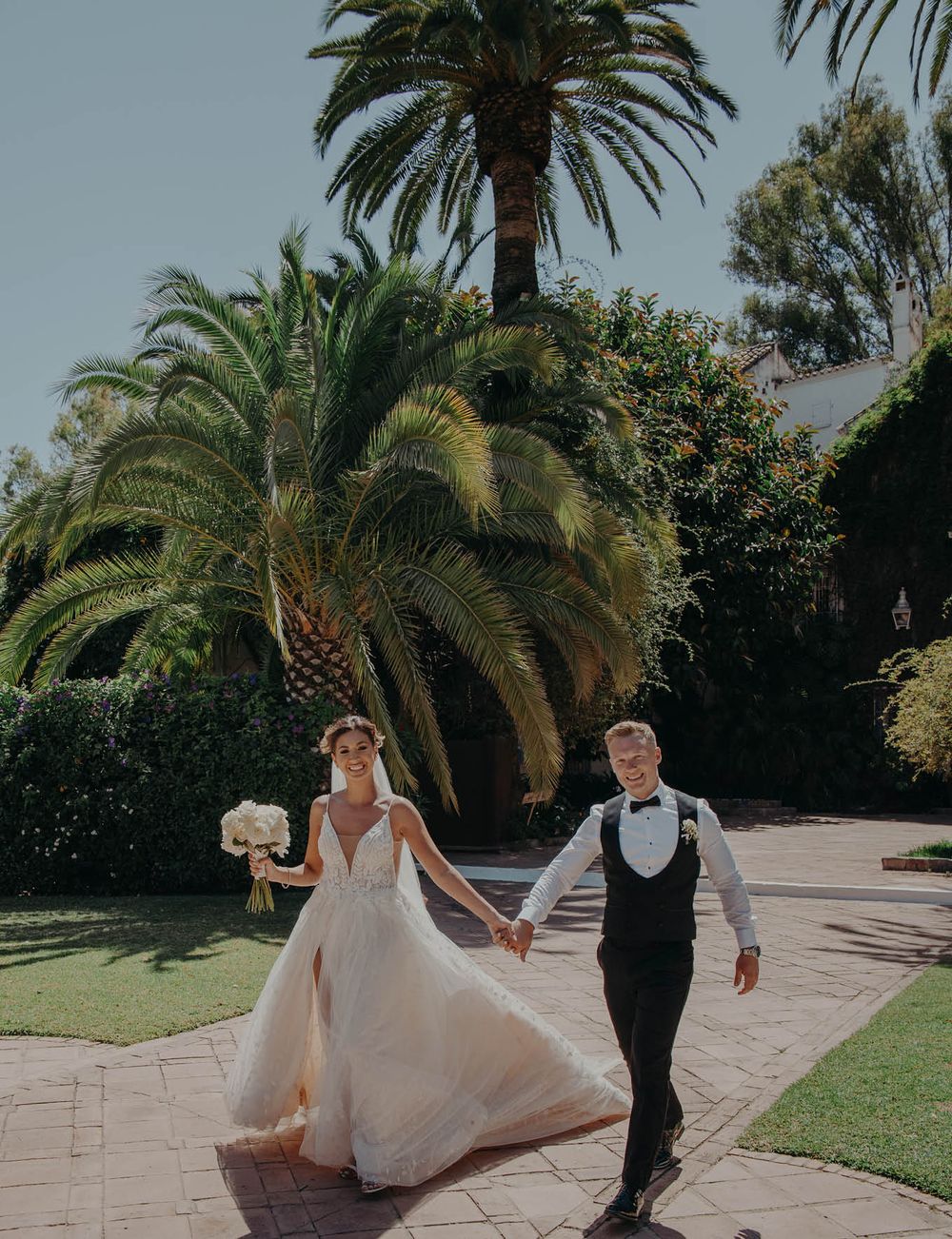 A romantic couple walks hand in hand through a tropical palm garden on their wedding day.