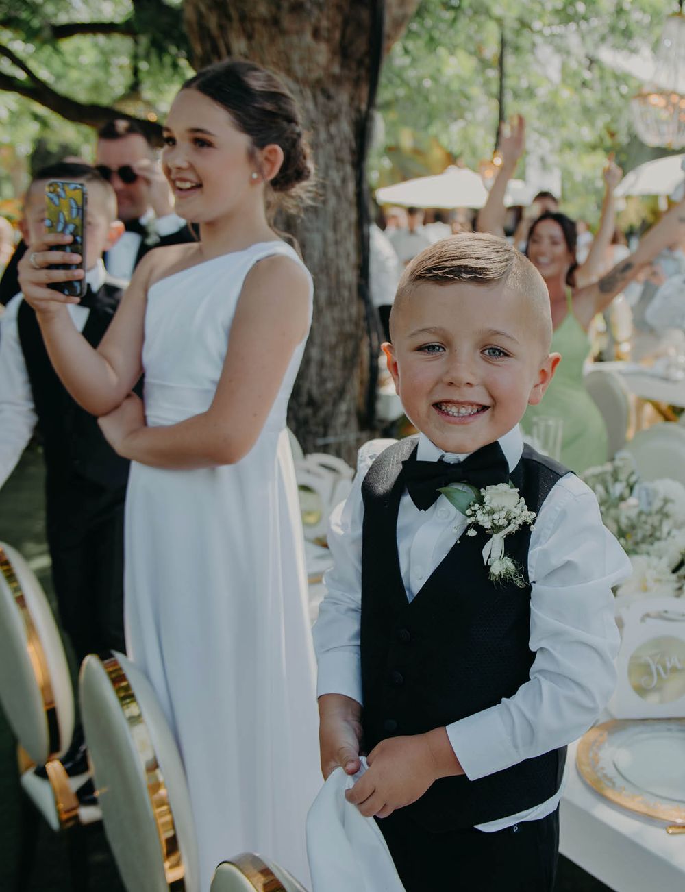 Young attendants in formal wedding attire smile during an outdoor reception celebration.