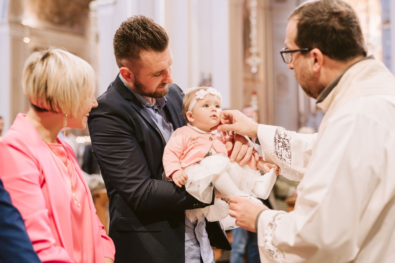 Bambina in chiesa riceve uno dei riti del Battesimo dal prete mentre il papà la tiene in braccio e la mamma le sorride accanto.