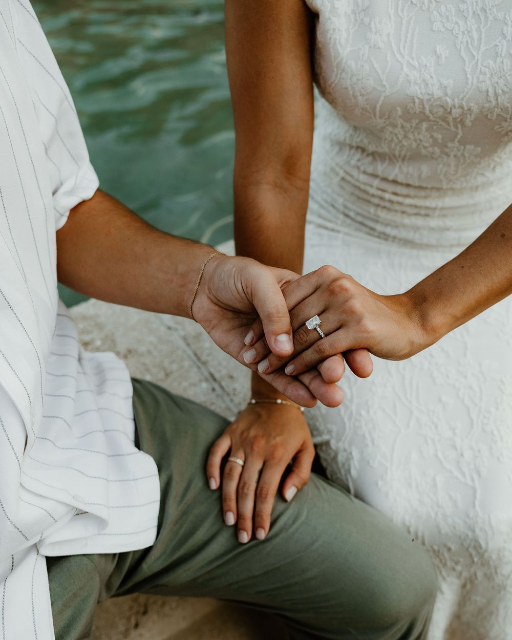 Close-up details of hands with wedding rings and white dress fabric against a water backdrop.