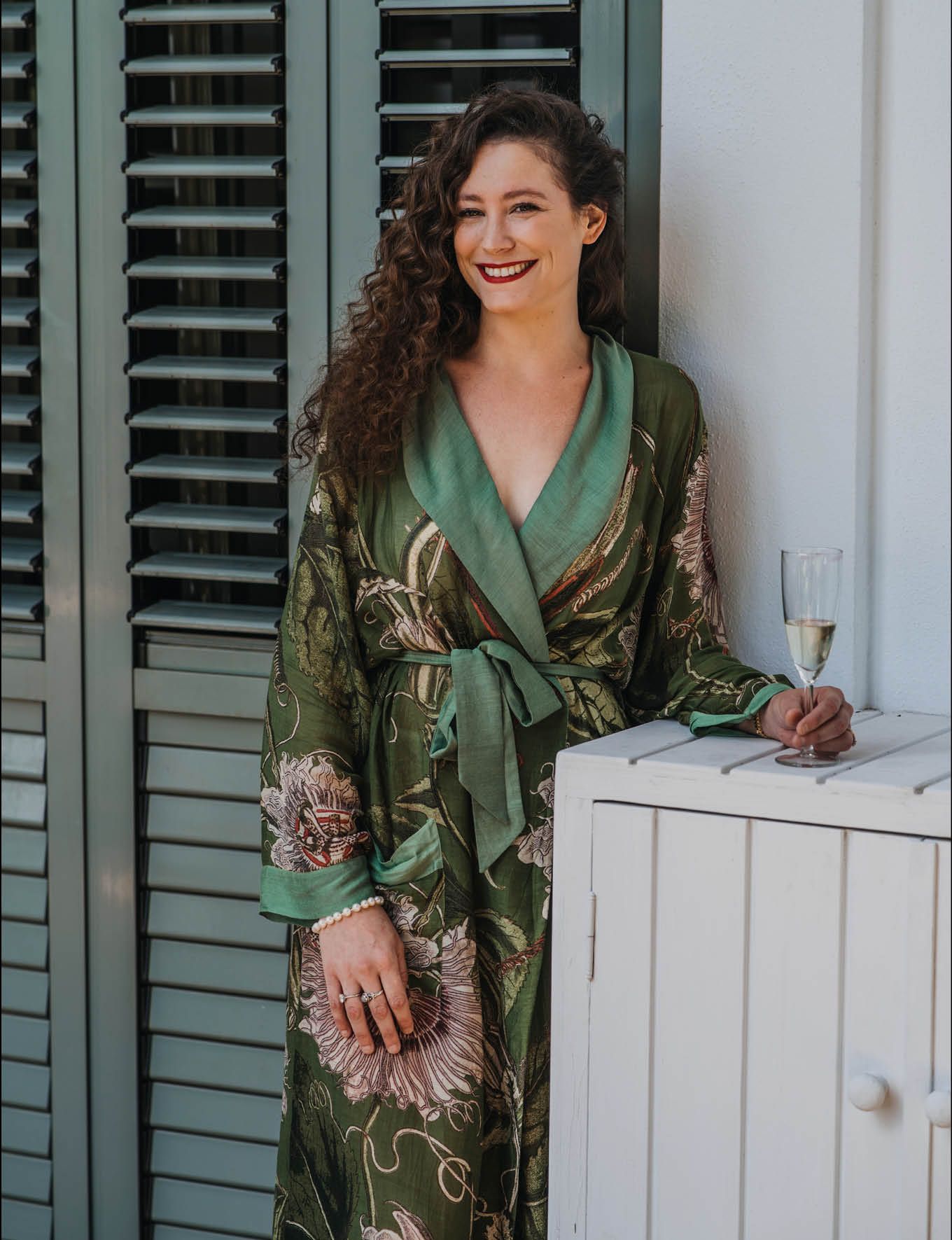 Person in floral green silk robe holding champagne glass against shuttered backdrop.
