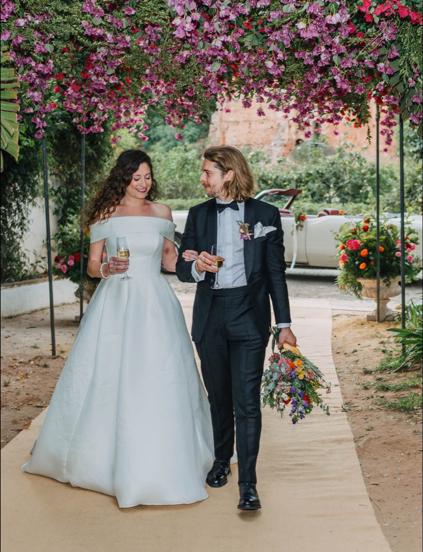 A wedding couple strolls beneath pink floral archway on their special day.