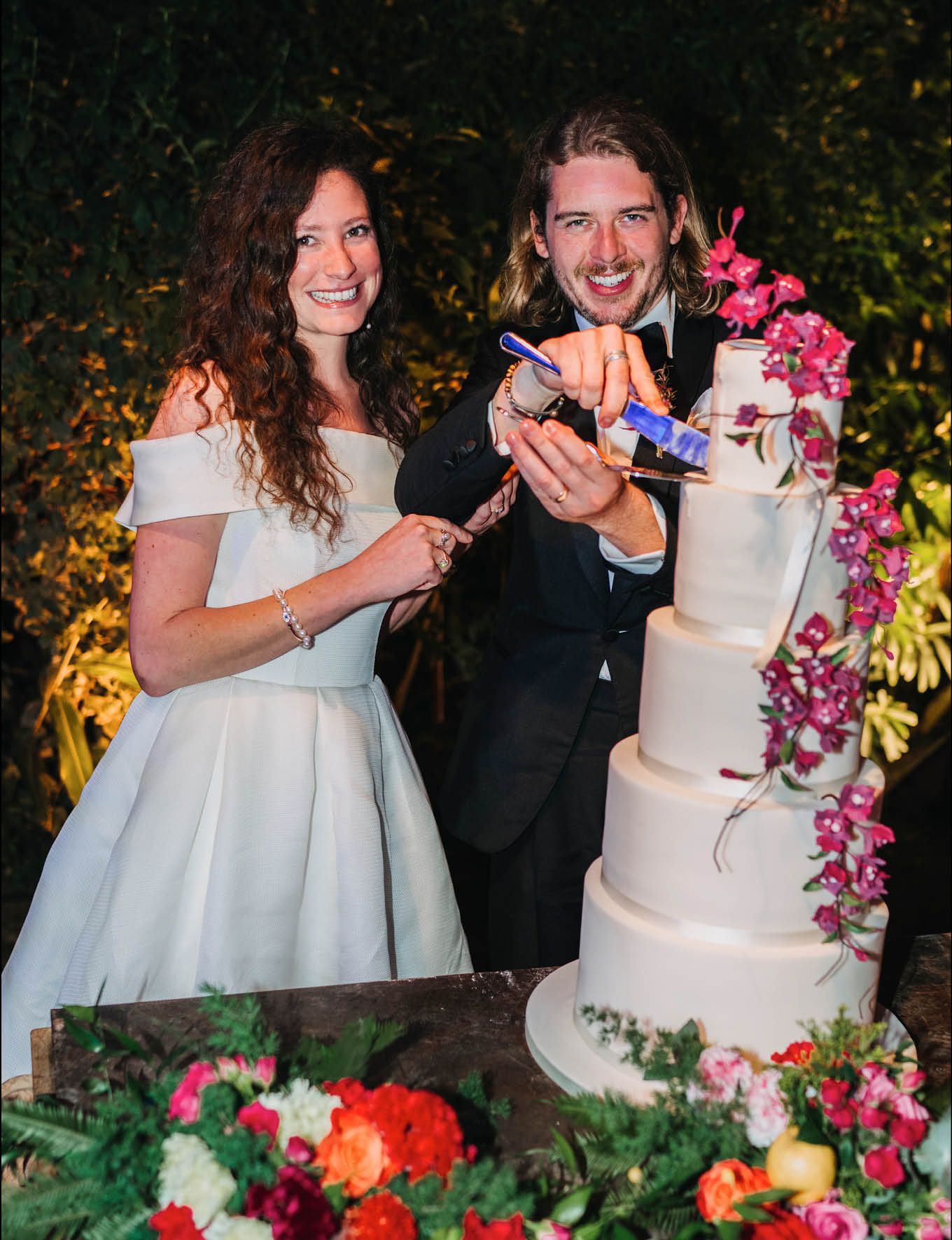 Wedding couple cuts into their elegant white tiered cake decorated with pink orchids.