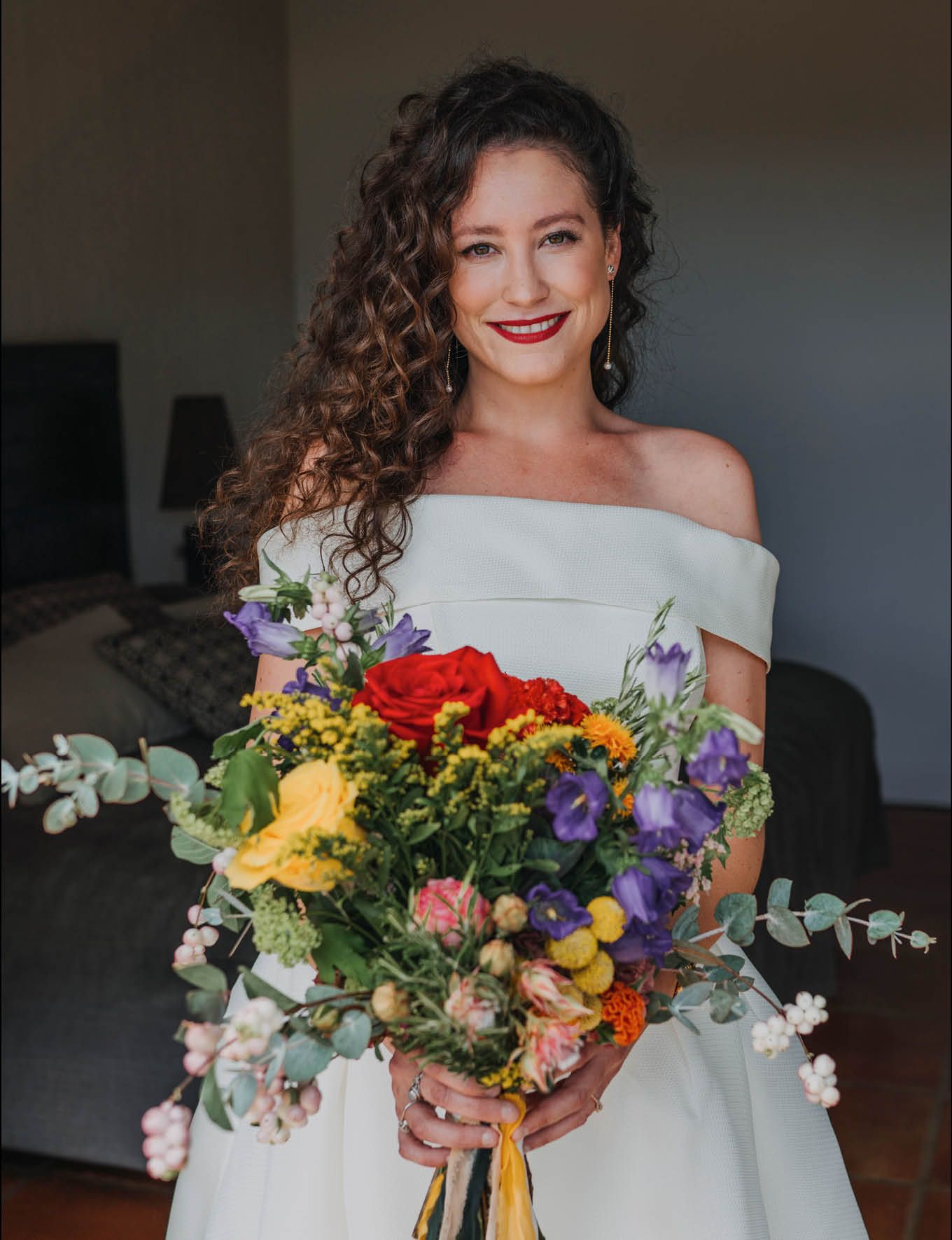 Off-shoulder bridal portrait with vibrant wildflower bouquet featuring red roses and purple blooms.