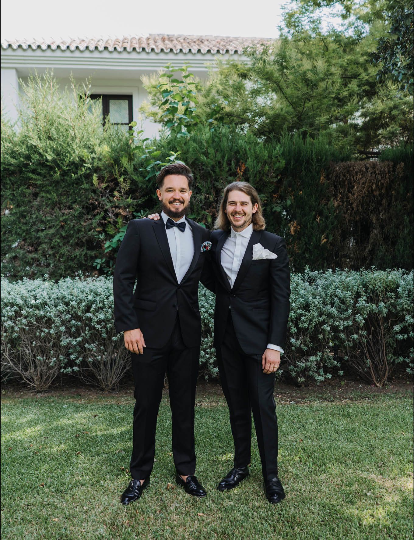 Wedding party members in black tuxedos posing in garden setting.