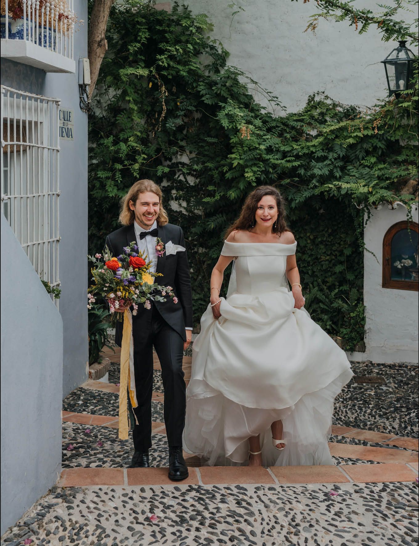 Wedding couple walking on cobblestone path with colorful bouquet and flowing white dress.