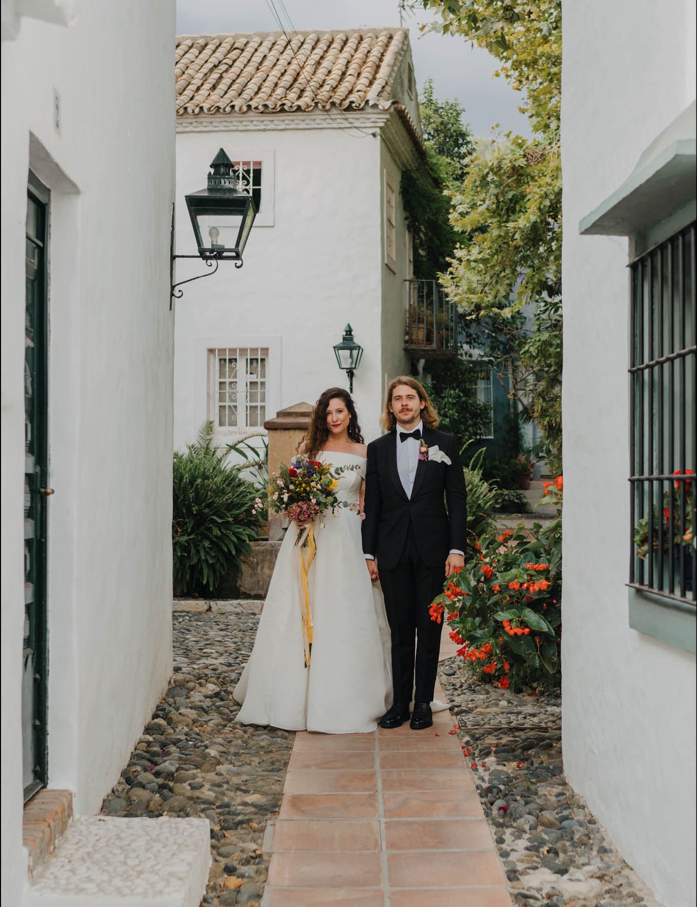 Newlyweds posing in Mediterranean-style courtyard with white walls and vintage lanterns.