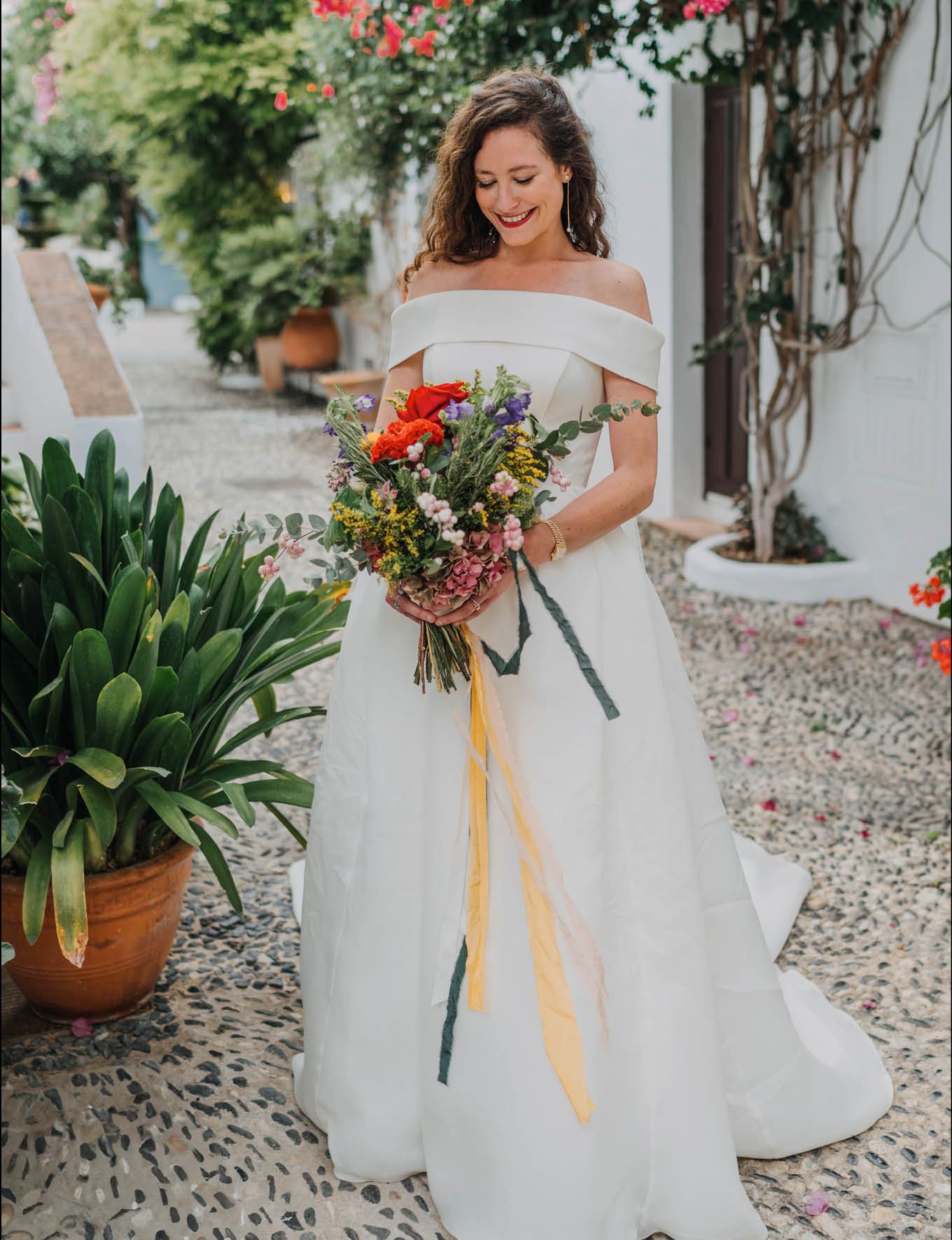 Bride in off-shoulder gown holding colorful bouquet with yellow ribbons in garden setting.