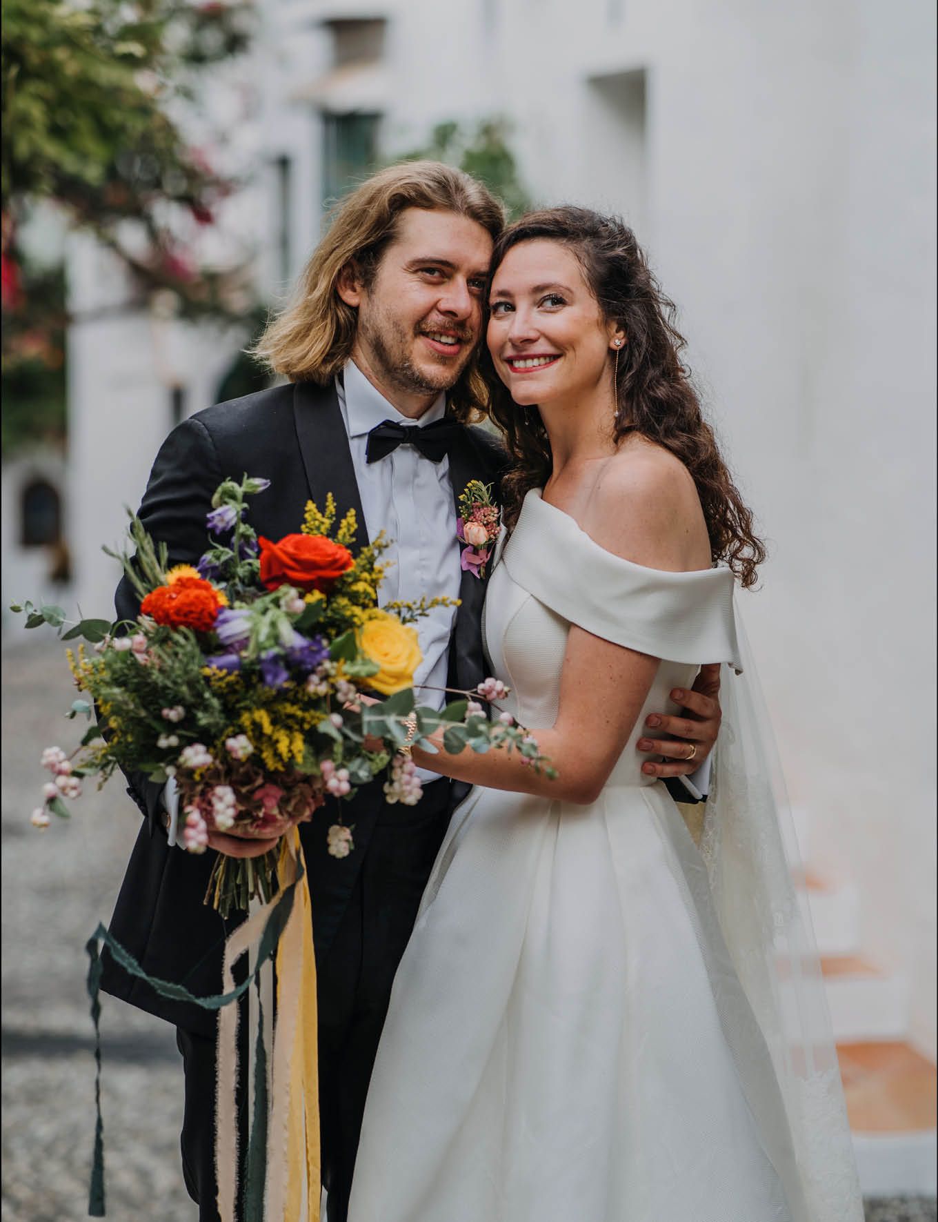 Wedding couple sharing joyful moment with vibrant bouquet against white wall.