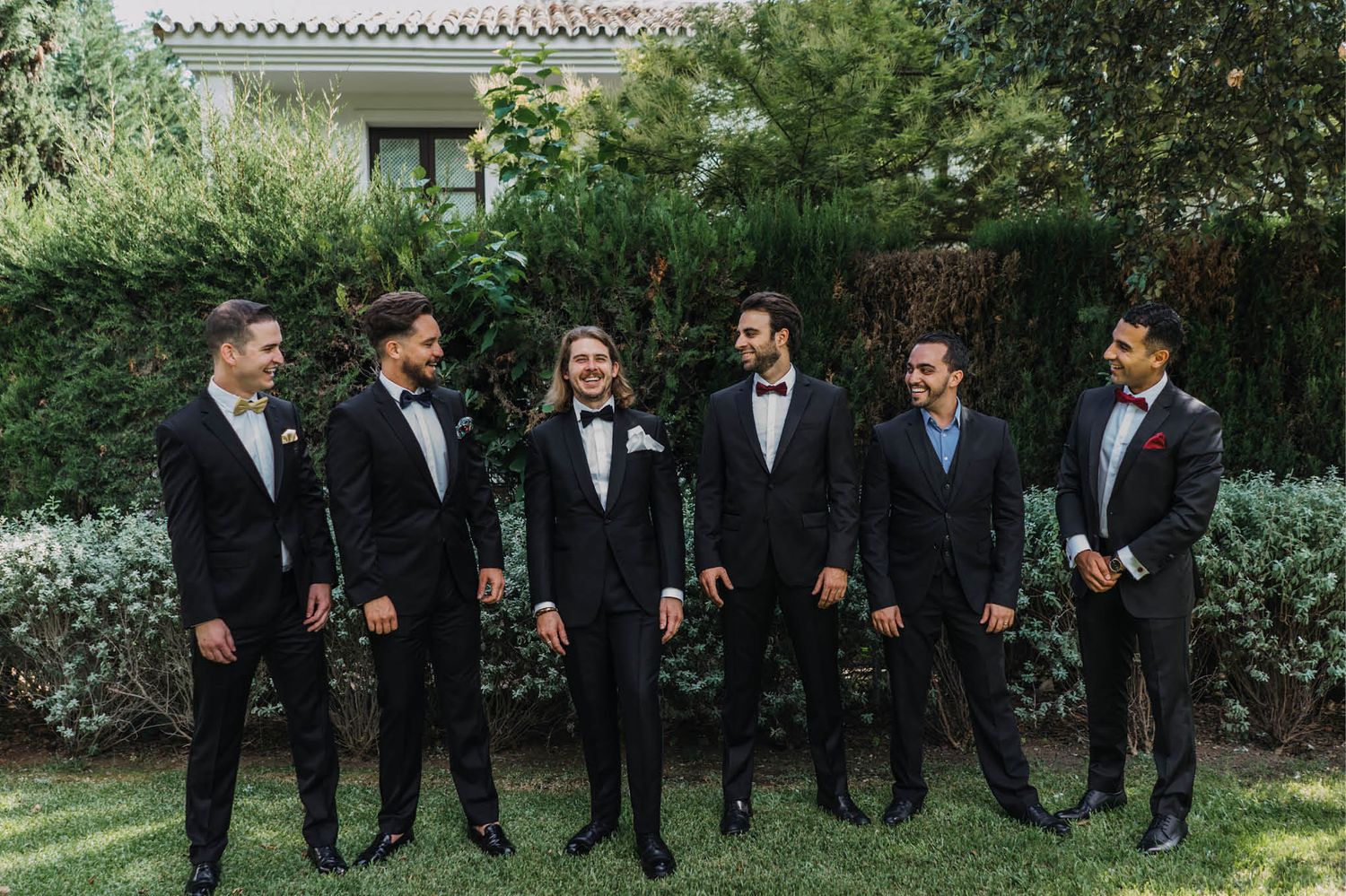 Groomsmen in black tuxedos and bow ties pose together in a garden setting.