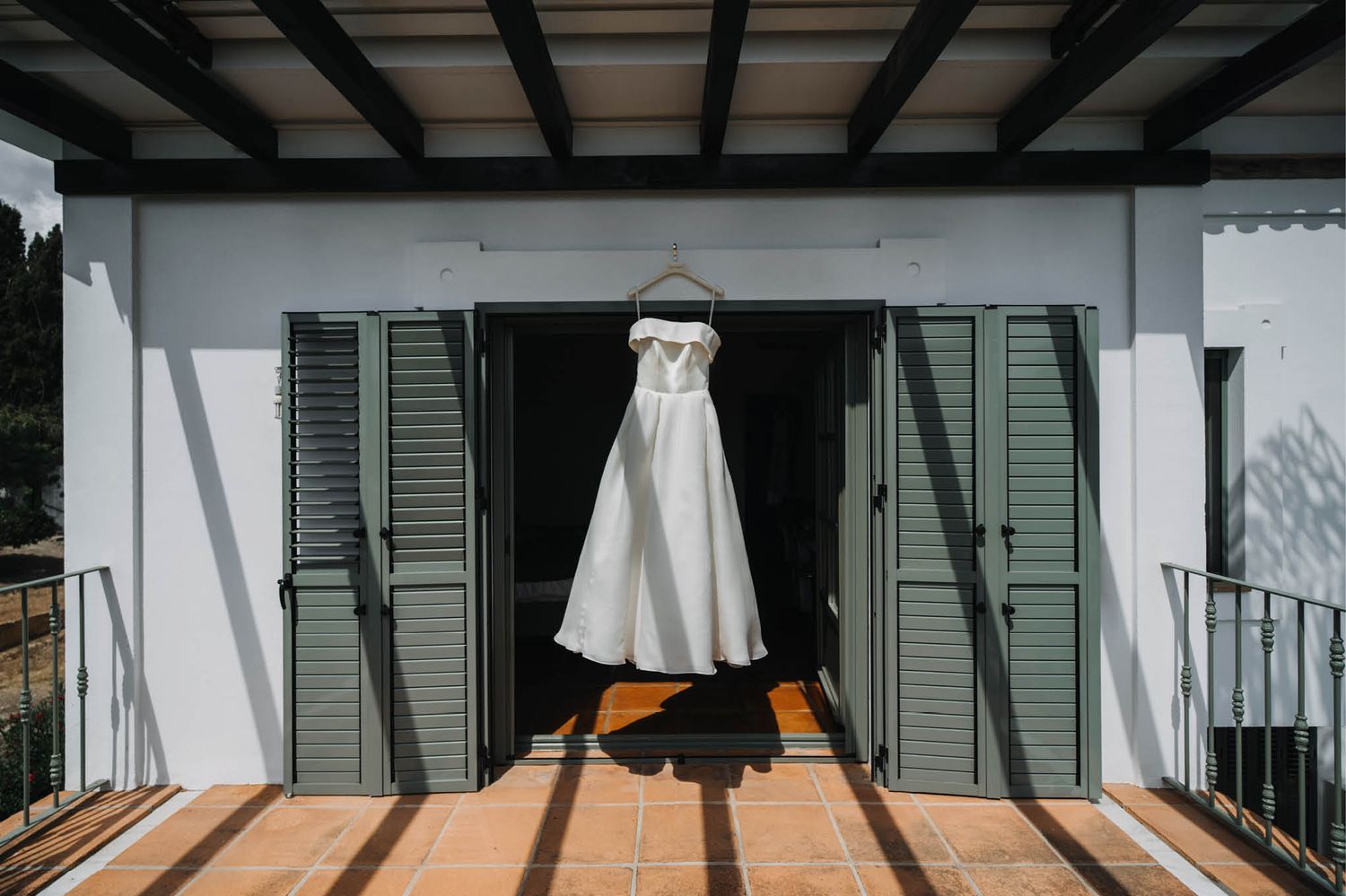 Wedding dress hangs dramatically between green shutters on white villa balcony.