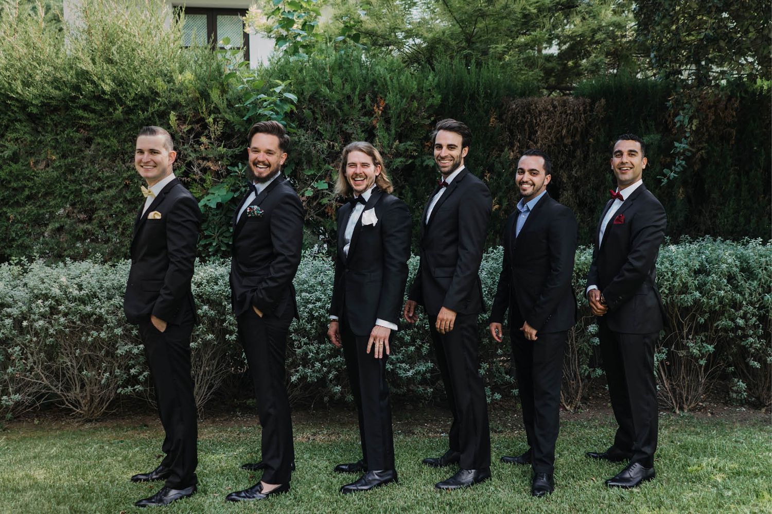 Wedding party lined up outdoors wearing formal black suits against garden backdrop.