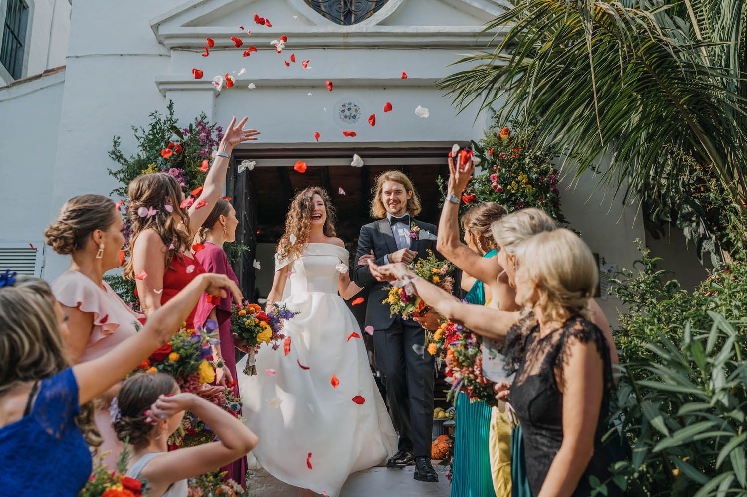 Wedding party throws red rose petals outside a white building entrance.