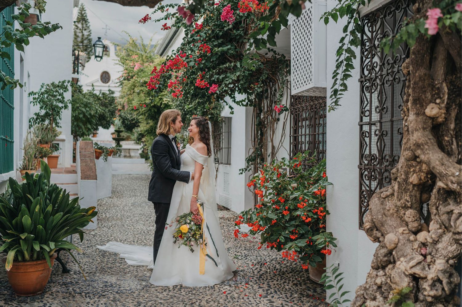 A romantic wedding couple poses in a Mediterranean courtyard with climbing red flowers and white stucco walls.