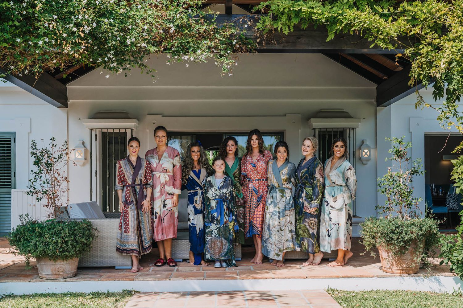 Group wearing floral and patterned robes gathered on a white porch with greenery overhead.