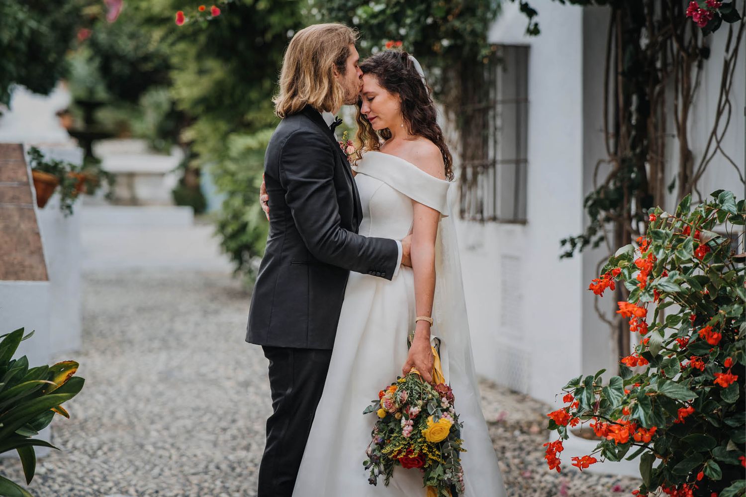 A romantic moment shared between newlyweds along a cobblestone path with vibrant climbing flowers.