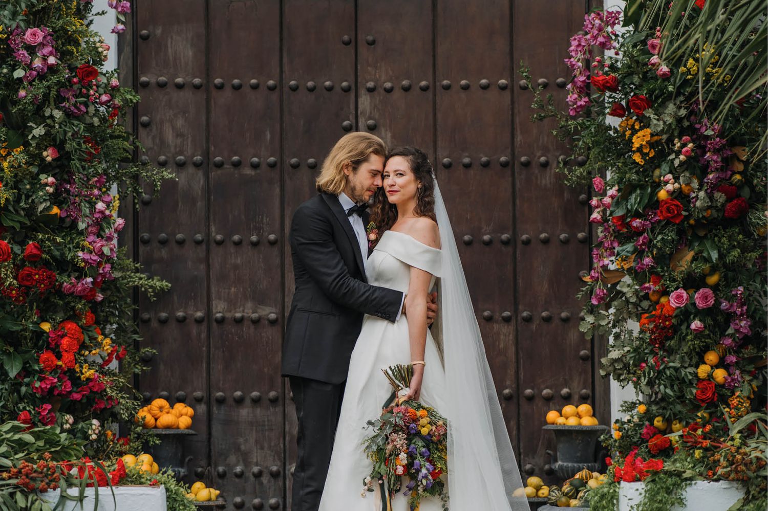 Romantic wedding portrait against a rustic wooden door adorned with colorful floral arrangements.