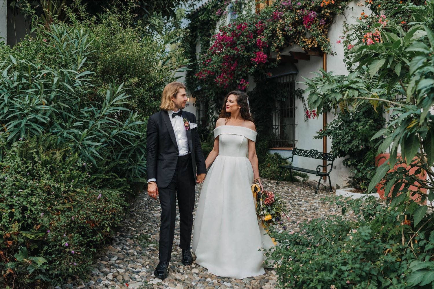 Elegant wedding couple walking through a lush garden pathway with Mediterranean architecture.