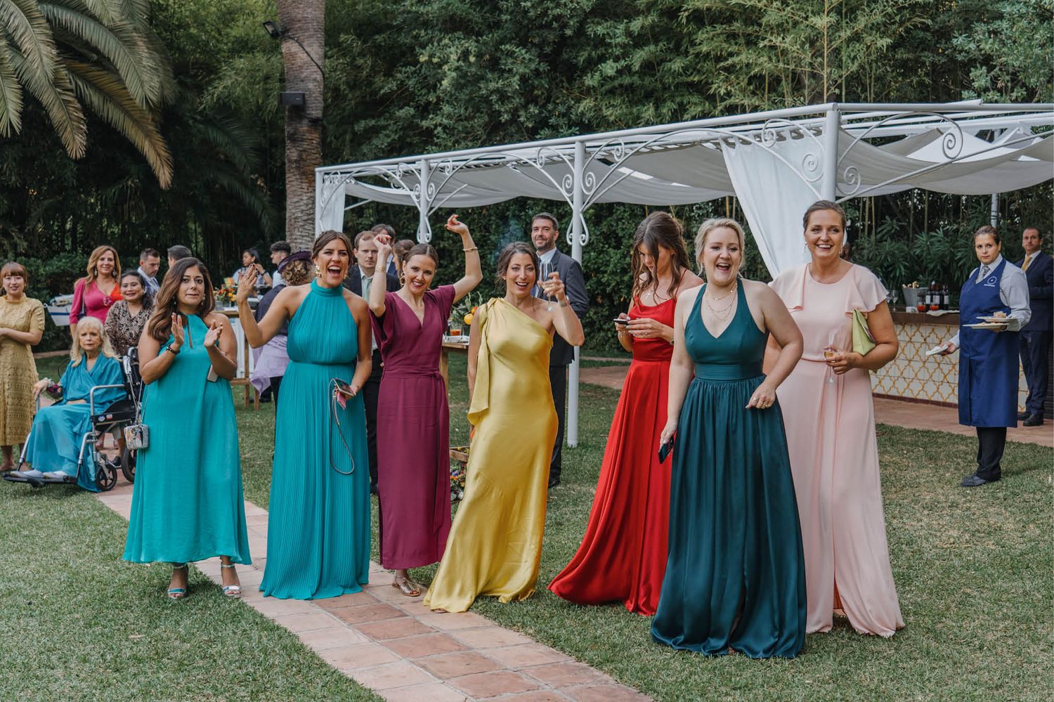 Colorful bridal party wearing rainbow-hued dresses standing in formation on a garden path.