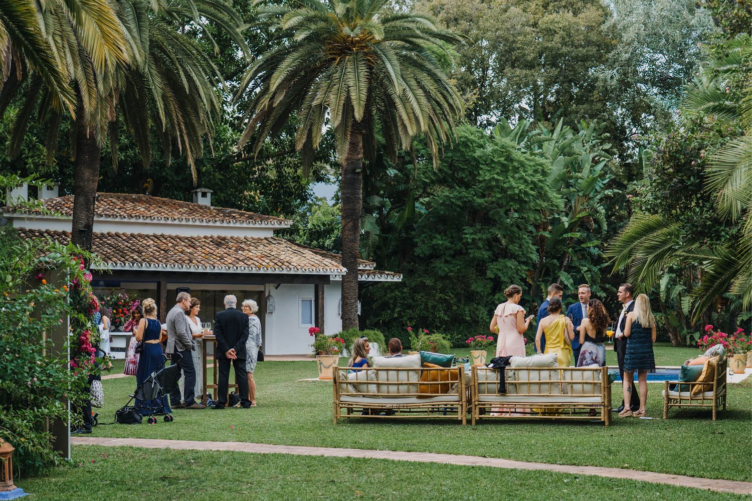 Guests gathering on a lush garden lawn with palm trees and a small white building during an outdoor event.