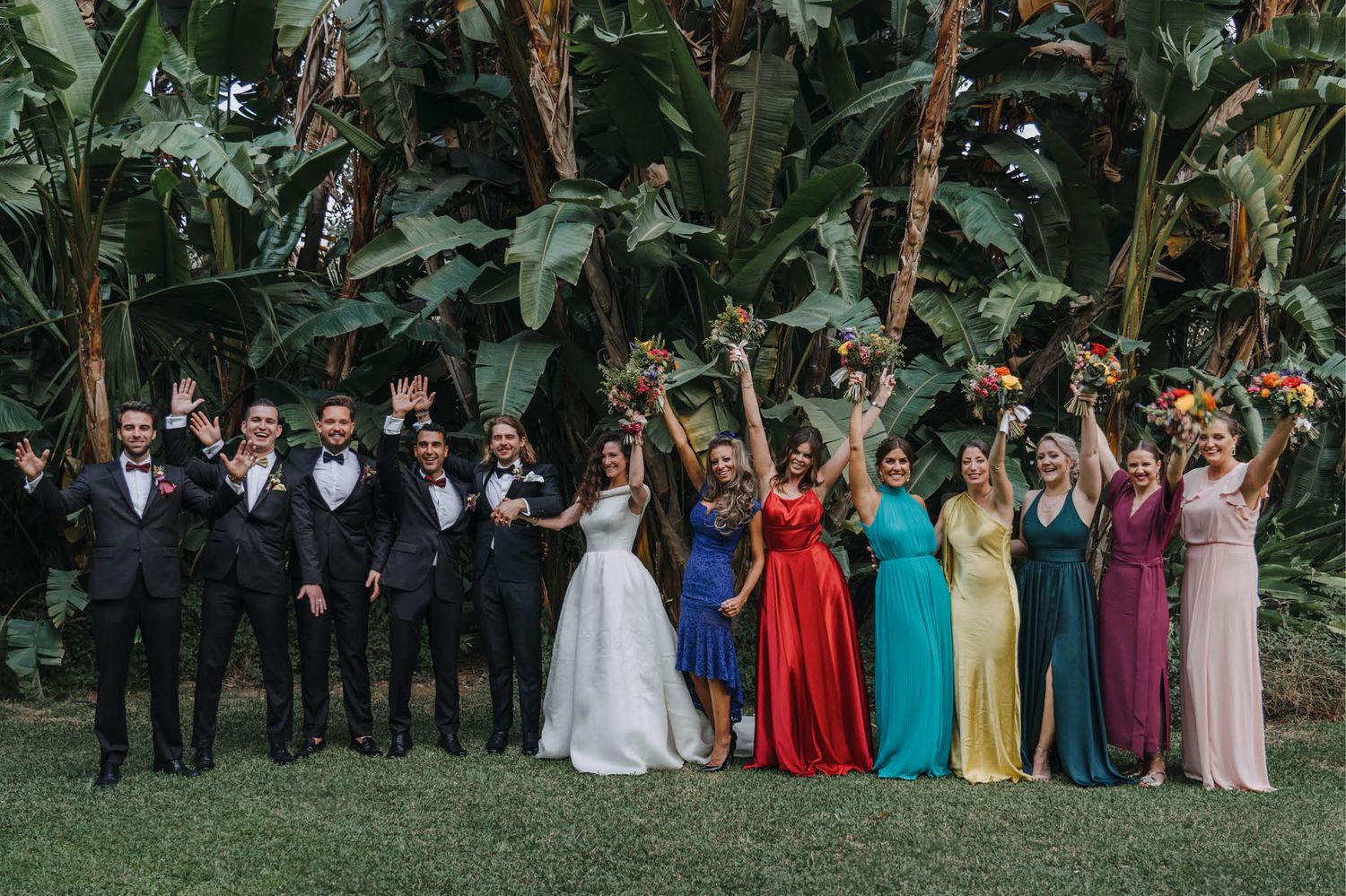 Wedding party lined up in formal attire against a backdrop of tropical plants.