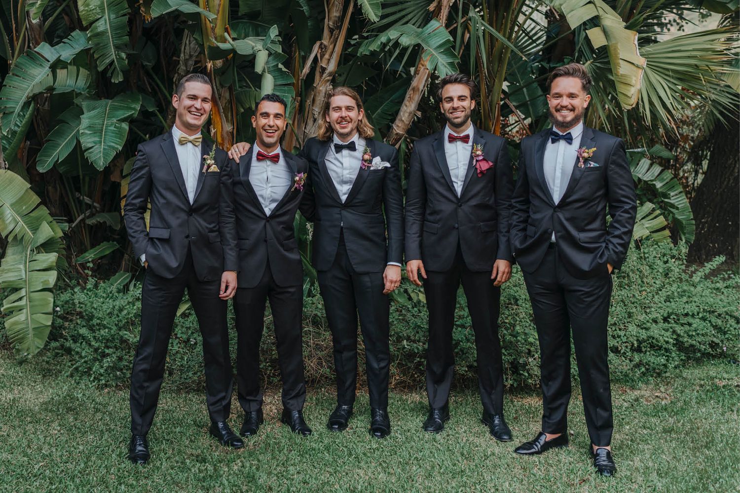 Groomsmen in black tuxedos posing together in a tropical garden setting.