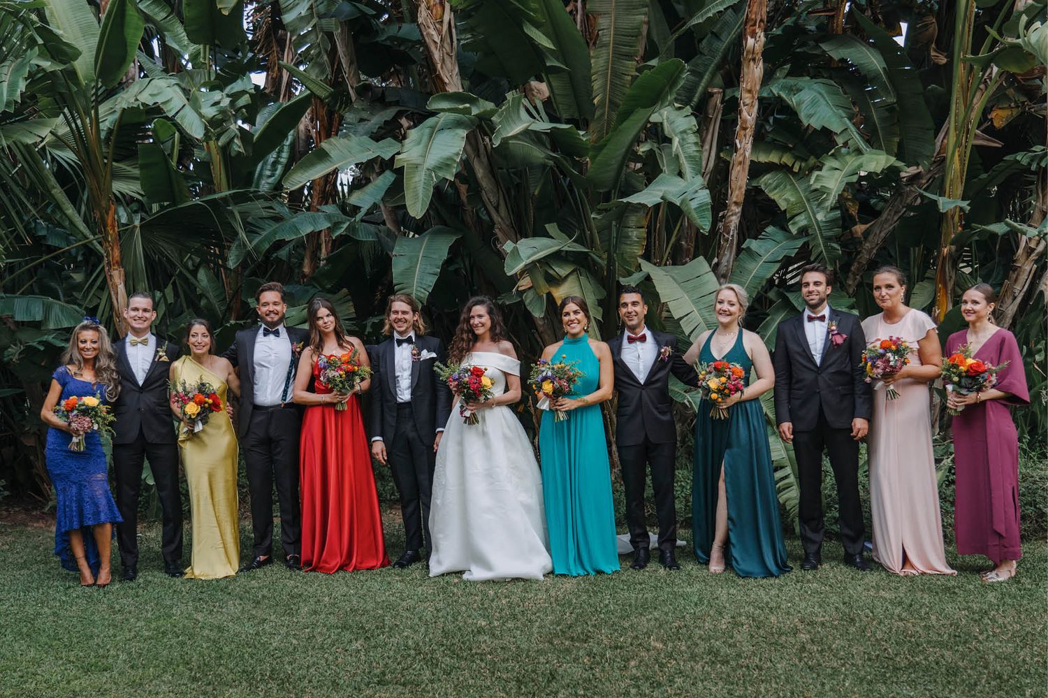 Wedding party in colorful bridesmaid dresses stands together on a lawn with tropical foliage backdrop.