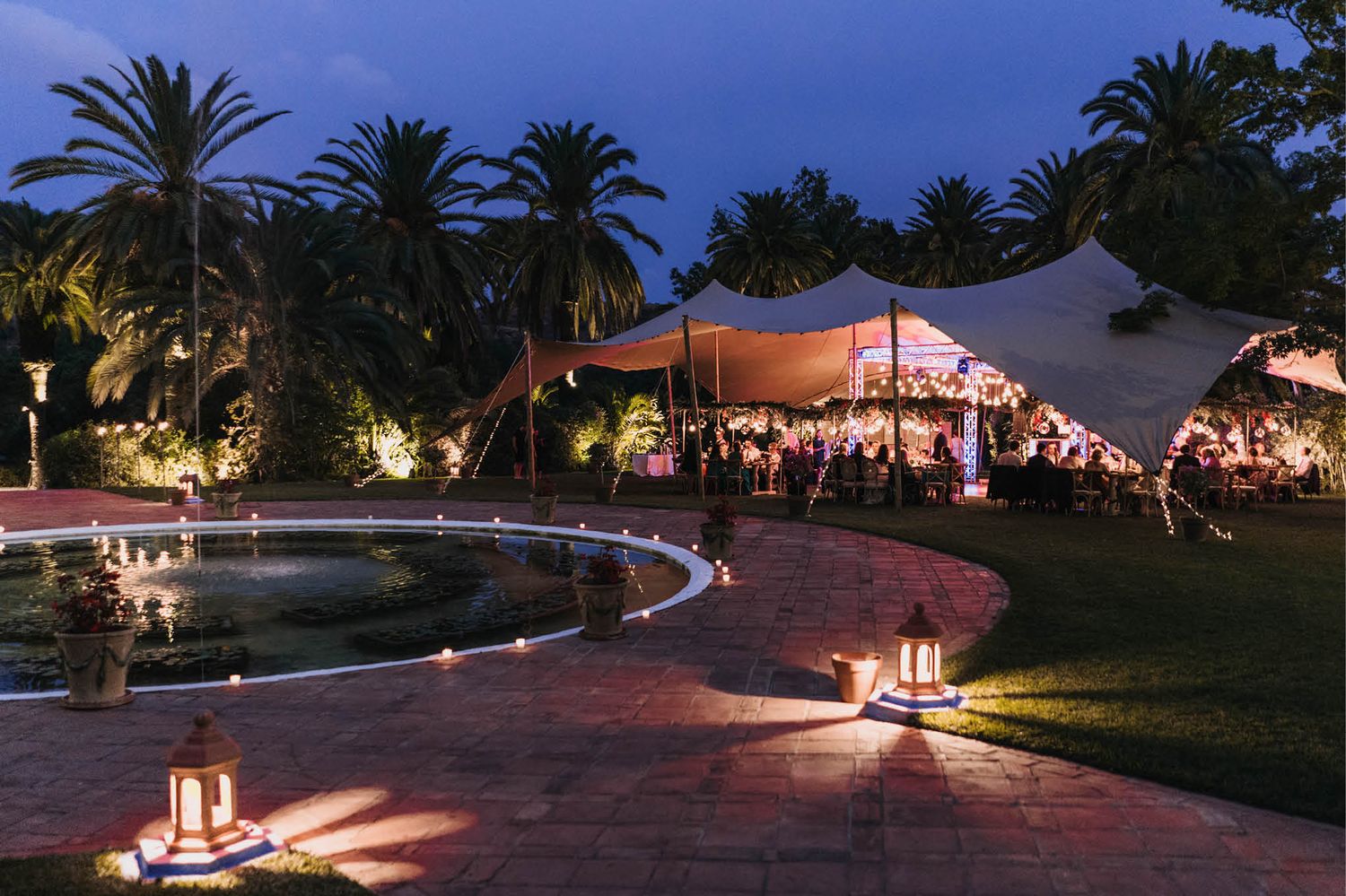 Evening outdoor reception with illuminated tent and lantern-lit pathway around a reflective pool.