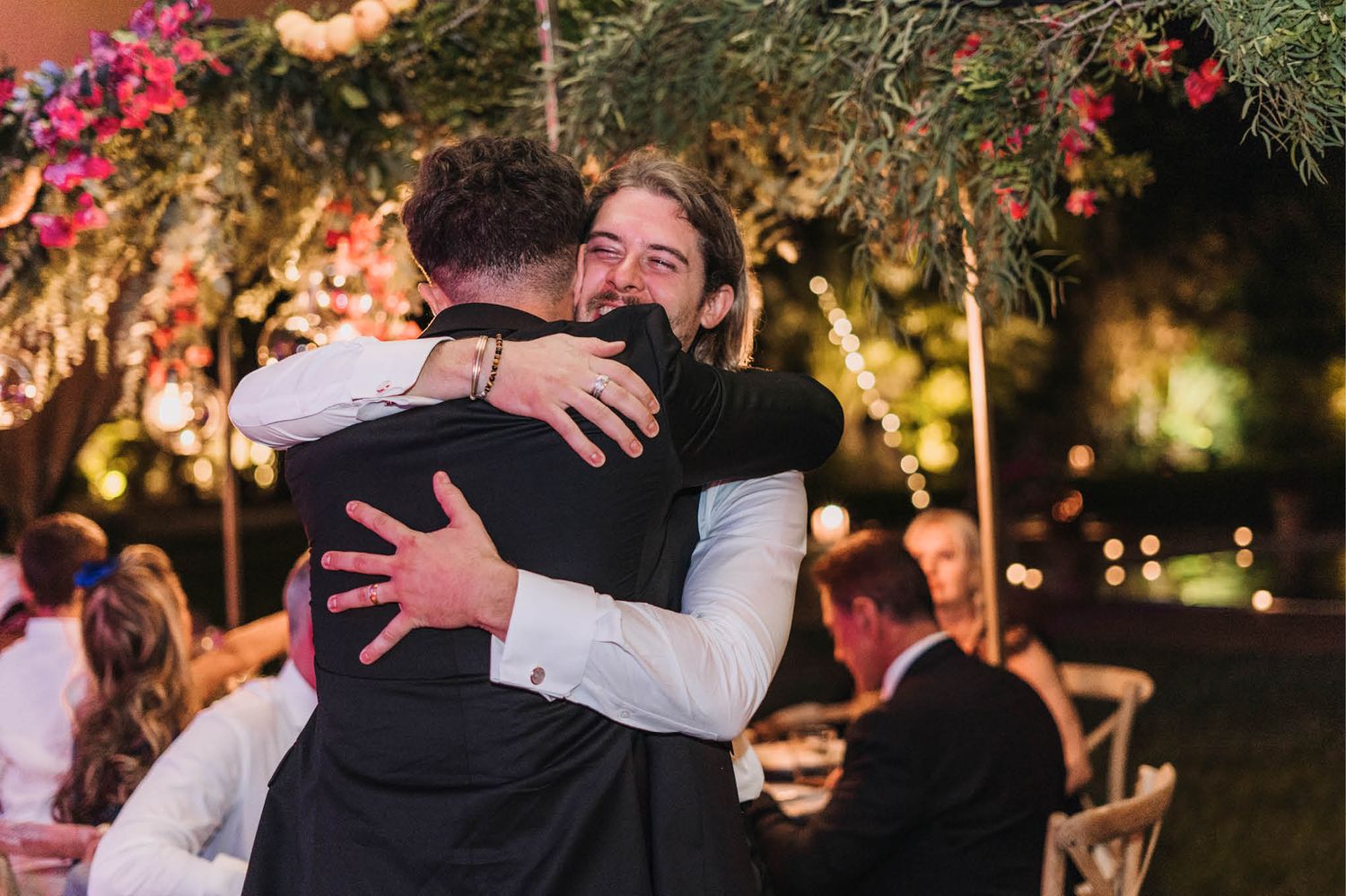 Emotional embrace during evening wedding celebration under string lights and floral decor.