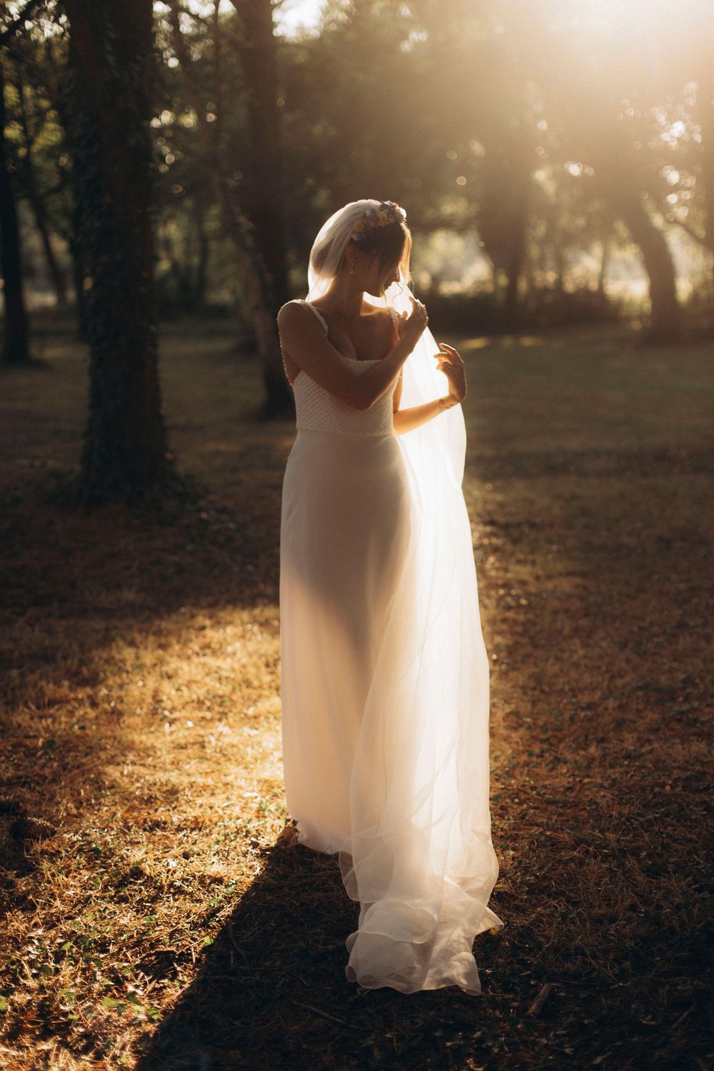 A bride in a white wedding dress stands in golden sunlight filtering through autumn trees in a park setting.